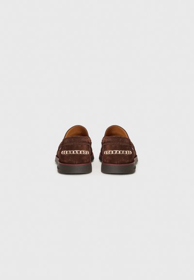 Mocassins en suède marron avec un accent en chaîne, en forme arrondie et avec une semelle plate. Texture lisse avec des détails de couture visibles le long des bords.