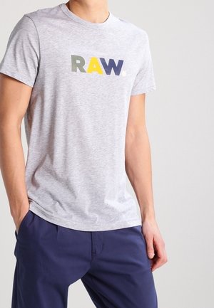 Personne portant un t-shirt gris avec "RAW." imprimé en lettres vertes, jaunes et bleu foncé, associé à un pantalon bleu marine.