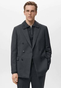Blazer doppiopetto grigio scuro con colletto nero testurizzato e dettagli sottili sulle tasche, indossato su un maglione scuro a maglia.