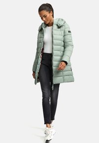 Abrigo tipo puffer verde claro con costuras horizontales, capucha desmontable y cremallera frontal. Llevado sobre una camiseta blanca y jeans negros.