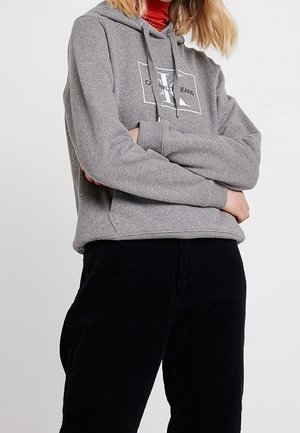 Personne portant un sweat à capuche gris Calvin Klein Jeans par-dessus un col roulé rouge et un pantalon noir, debout les bras croisés devant un fond uni.