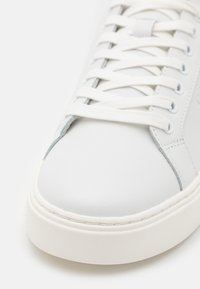 Sneaker in pelle bianca con punta tonda, dettagli testurizzati e accenti grigi. Presenta lacci piatti bianchi e una spessa suola in gomma bianca.