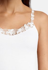 White tank top with lace trim on straps and neckline, featuring floral patterns and a smooth texture. Simple design and fitted shape.