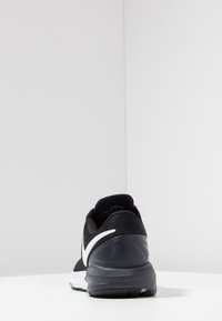 Scarpa da ginnastica nera con parte superiore in mesh testurizzato, accento bianco a forma di swoosh e suola in gomma grigia, dotata di tacco imbottito e punta arrotondata.