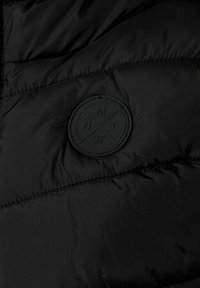Svart puffjacka med quiltad textur. Har en rund, präglad svart patch med "NMT" och "97" på. Enkel design.