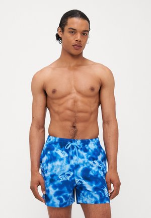 Blå tie-dye badebukser med snor i livet, med en glat tekstur og gentagne hvide logoer over stoffet.