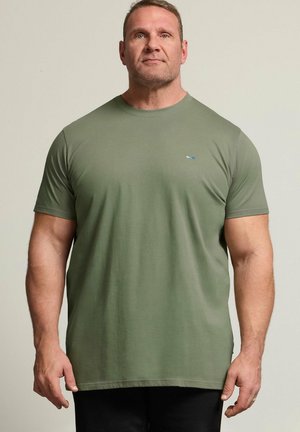 Homme musclé de face portant un t-shirt manches courtes uni vert olive et un pantalon noir sur un fond clair uni.