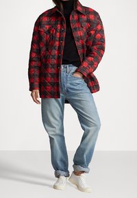 Giacca a diamante rossa e nera con bottoni a pressione, abbinata a jeans azzurri con risvolti e sneakers bianche.