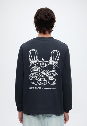 Persona que lleva una camiseta negra de manga larga con un gráfico blanco de una mesa de café, sillas, tazas de café y pasteles en la espalda.