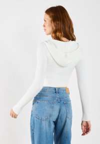 White cropped hoodie with ribbed texture, featuring a hood and long sleeves, paired with high-waisted blue denim jeans.