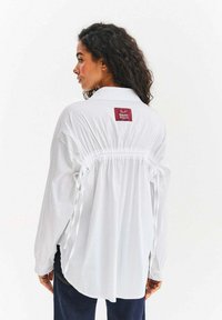 Biała koszula z długim rękawem z marszczonym tyłem, regulowanymi troczkami i burgundową metką z logo. Gładka tkanina, luźny krój i zaokrąglony dół.