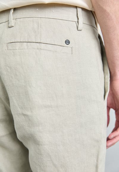 Pantalons en lin beige clair avec une finition texturée, présentant une poche arrière et un petit détail de bouton logo noir. Design ajusté.