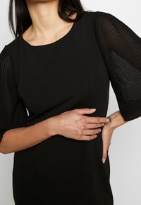 Czarna sukienka z gładkiego materiału, z teksturowanymi plisowanymi rękawami i okrągłym dekoltem. Dopasowany krój z subtelnymi szwami bocznymi.