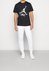 Muž v černém grafickém tričku Nike Flight, bílých teplácích a bílých teniskách stojí proti jednoduché bílé zdi.