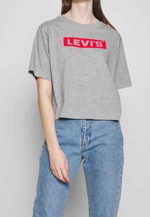 Persoon draagt een grijs Levi's T-shirt met rood logo en lichtblauwe high waist jeans, staand tegen een effen achtergrond.