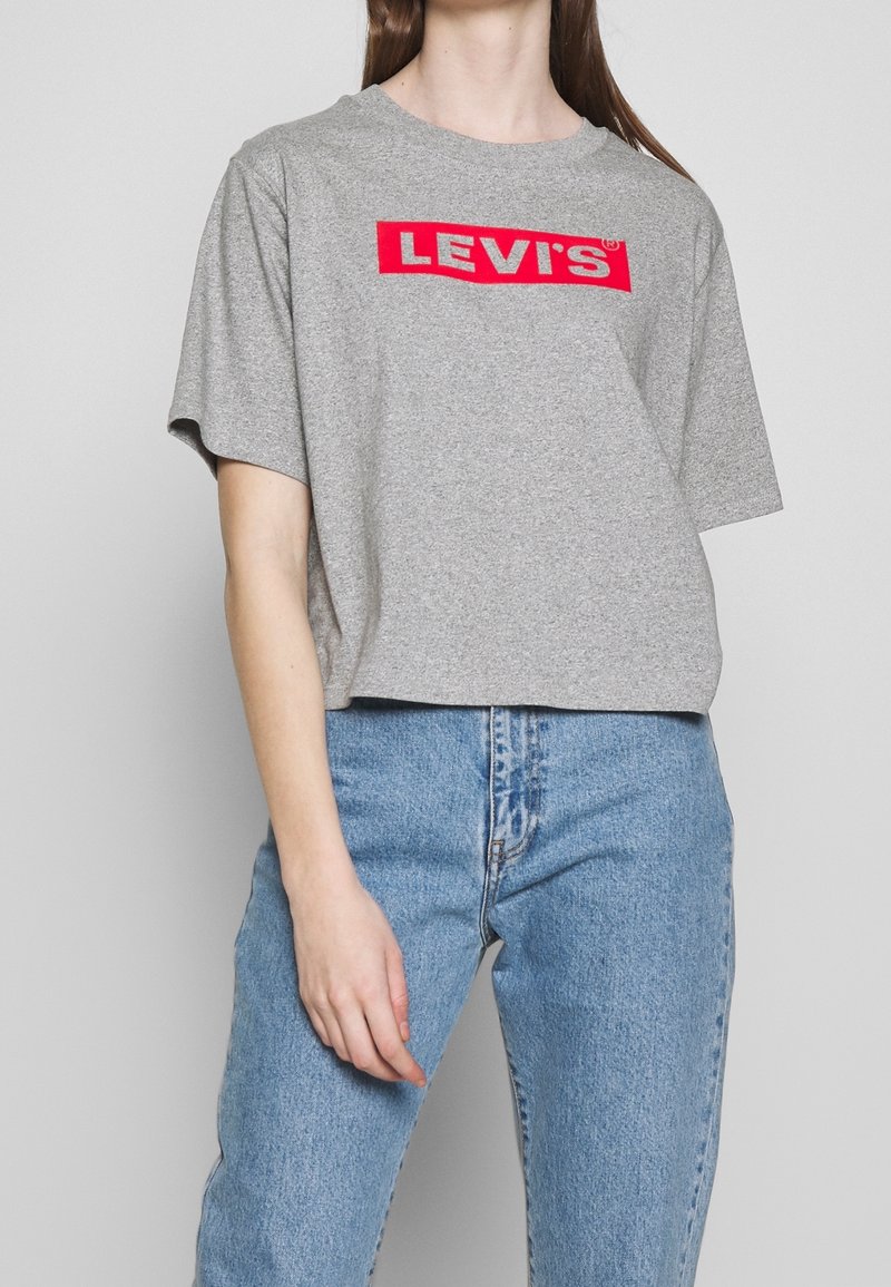 Personne portant un t-shirt gris Levi's avec logo rouge et un jean taille haute bleu clair, debout devant un fond uni.