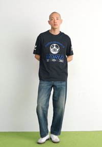Sort Adidas T-shirt med et blåt grafisk design, der inkluderer en fodbold og tekst, parret med lyseblå jeans og hvide sneakers.