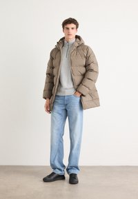 Pufferjack in taupe, met een capuchon en ritssluiting, gedragen over een grijze trui. Gecombineerd met lichtblauwe recht-toe-rechtdoor jeans en zwarte sneakers.