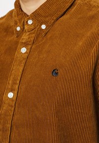 Camisa de pana marrón con cuello abotonado, botones blancos y un pequeño logo negro en el lado izquierdo. Textura de costillas verticales.