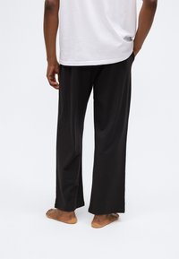 Pantalons noirs légers à jambes larges avec une texture lisse, présentant une coupe décontractée et un subtil détail d'ourlet, portés pieds nus.