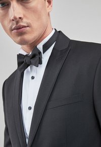 Schwarze Smokingjacke mit satinbesetzten Revers und einer einzigen Brusttasche, kombiniert mit einem weißen Button-Down-Hemd und einer schwarzen Fliege.