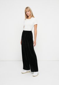 Jonge vrouw met stijlblond haar, gekleed in een witte top met korte mouwen, zwarte wijde broek en witte sneakers, staande op een witte achtergrond.