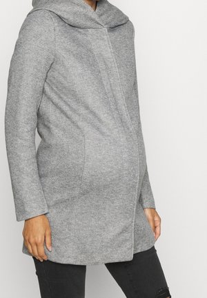 Vrouw draagt een nauwsluitende grijze zwangerschapsjas met een hoge kraag en zwarte gescheurde jeans, staand tegen een effen witte achtergrond.