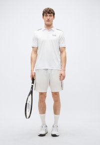 Biela polokošeľa s čiernymi akcentmi a logom, v kombinácii s ľahkými šortkami. Stojí s tenisovou raketou, nosí biele ponožky a topánky.