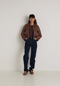 Jaqueta de couro marrom com zíper, blusa ajustada marrom, jeans de cintura alta azul escuro, sapatos marrons com padrão e cadarços laranja, boné preto.