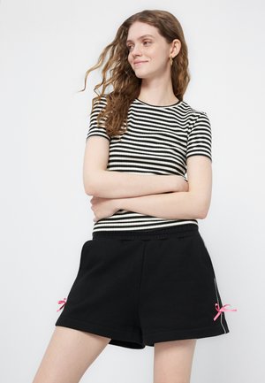 Ung kvinde med bølget hår iført sort-hvid stribet kortærmet top og sorte shorts med lyserøde bånddetaljer, armene krydset, smilende.