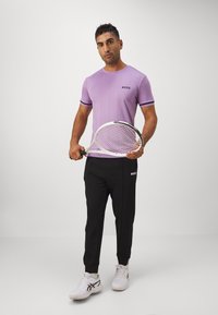 Lila Sport-T-Shirt mit schwarzen Akzenten und Logo, kombiniert mit schwarzen Hosen und weißen Sneakers, während er einen Tennisschläger hält.