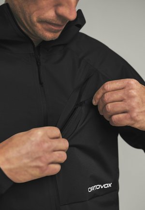 Mann trägt eine schwarze Ortovox-Jacke und öffnet eine kleine Brusttasche auf der linken Seite.