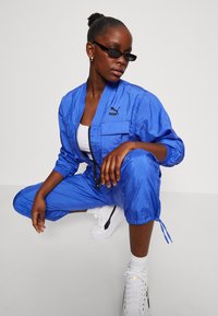 Chándal ligero azul con cremallera frontal, puños elásticos y dos bolsillos; combinado con zapatillas blancas con detalles en negro. Lleva puestas gafas de sol oscuras.