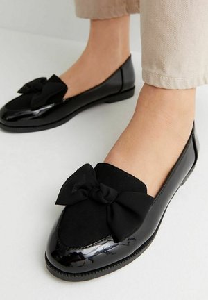 Black flat shoes with a glossy patent finish and a textured black bow detail on the front. Featuring a rounded toe and low outsole.