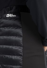Zwarte gewatteerde vest met een gladde textuur, met logo-details en een contrasterend materiaal van een effen zwarte stof aan de zijkant.