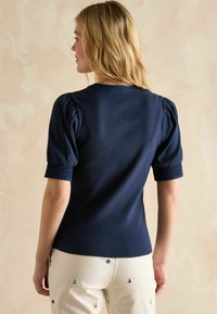 Tumedat sinise ribadega pluus lühikeste puhvärvidega ja keha lähedase disainiga. Omab ripsmetega mansette ja ümmargust kaelust, koos heledate pükstega.