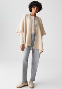 Beige gestrickter Poncho mit geripptem Muster, offene Vorderseite, getragen über einem hellen Hemd mit Knöpfen. Das Modell kombiniert ihn mit hellgrauen Jeans und beigen Sneakers.