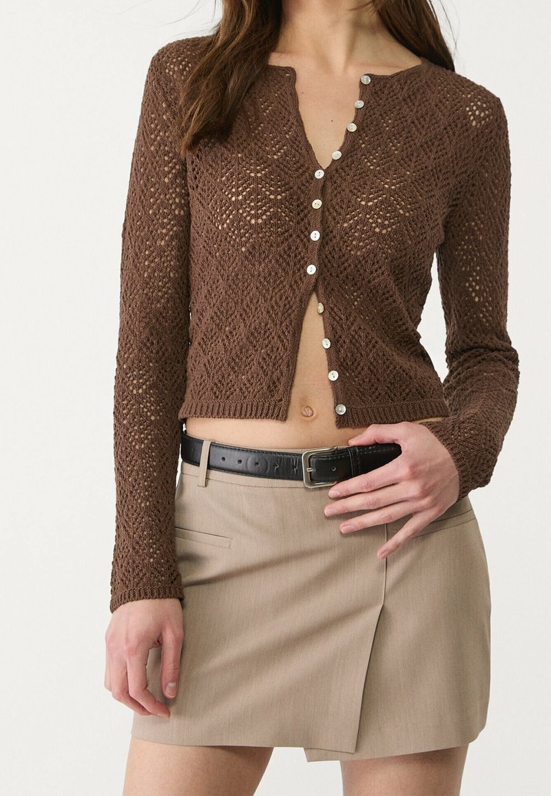 Cardigan lavorato a maglia marrone con motivo a diamante, maniche lunghe e bottoni argentati sul davanti. Abbinato a una gonna beige e una cintura nera.