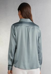 Camisa de satén de manga larga en un tono verde azulado suave, con una textura lisa, un cuello clásico y un detalle de plisado en la parte posterior.