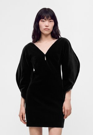 Femme aux longs cheveux noirs portant une robe en velours noire avec des manches longues bouffantes et un décolleté en V profond sur un fond blanc uni.