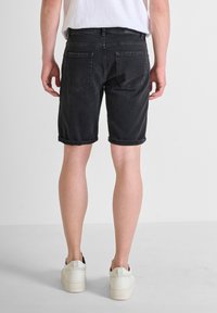 Shorts de mezclilla negra con dobladillos enrollados, dos bolsillos traseros y costuras de logo sutiles. Usados con zapatillas blancas sobre un fondo liso.