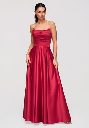 Vrouw draagt een rode satijnen strapless galajurk tot aan de vloer met een geplooid lijfje, poseert met de handen bij de zijkanten van de jurk en kijkt naar links.