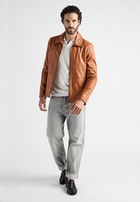 Brun læderjakke med stående krave, parret med en lys grå sweater og lys grå opsmøgte jeans, der har sorte sko.