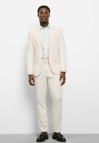 Hellbeiger Anzug mit tailliertem Schnitt, bestehend aus einem einreihigen Sakko und maßgeschneiderten Hosen. Kombiniert mit einem blau gestreiften Hemd und dunklen Schuhen.