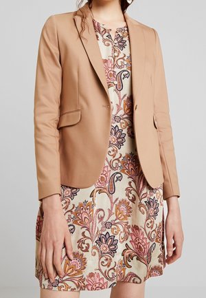 Kobieta ubrana w beżową dopasowaną marynarkę na kremowej sukience o długości do kolan z kwiatowym wzorem w kolorach różowym, pomarańczowym i fioletowym.