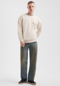 Lichtbeige sweatshirt met een ronde hals, voorzien van een borstzak. Gecombineerd met brede, vervaagde spijkerbroek en zwarte sneakers.