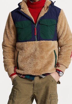 Person iført en beige fleecejakke med marineblå og grønne brystpaneler, rød rullekrave, ternet skjorte, khakifarvede cargobukser og armbåndsur.
