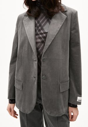 Femme portant un blazer en velours côtelé gris et un pantalon assorti par-dessus un col roulé noir et gris à motifs.