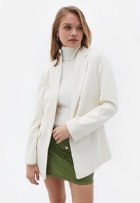 Blazer bianco su misura con rever a punta, abbinato a un dolcevita fitted color crema e una mini gonna verde, che presenta una texture liscia e linee pulite.
