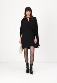 Črna mini obleka z značajem plašča, izrezanim ovratem in gladkim materialom. Ustrezno kombinirana z gležnjarji in svetlo obarvano torbico.